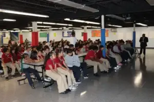 Students seated at cafeteria tables listen to a staff member during the assembly.