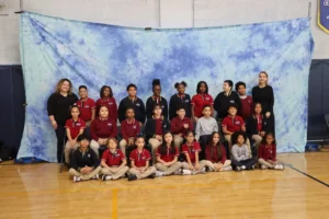 Two staff members stand with a class of students in maroon, red, and navy uniforms arranged in three rows in front of a blue tie-dye backdrop.