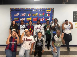 Eleven students in sports jerseys pose in two rows, several flashing peace signs, beneath a bulletin board reading "Different Cultures ONE Team" with flags from around the world.