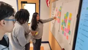 Students gather at a whiteboard covered in colorful sticky notes and a handwritten list of EntreX skills, as one points to a note and another holds a marker and a sticky note.