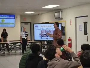 A presenter speaks enthusiastically beside a “Lion’s Hub: School-Based Access to Care” slide as a staff member stands nearby and students listen from their table.