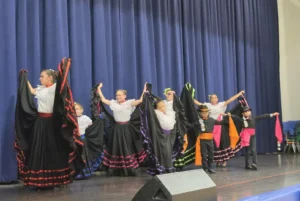 Girls in white blouses spread wide black skirts trimmed with colorful ribbons as two young boys in black hats hold out bright sashes in front of them.